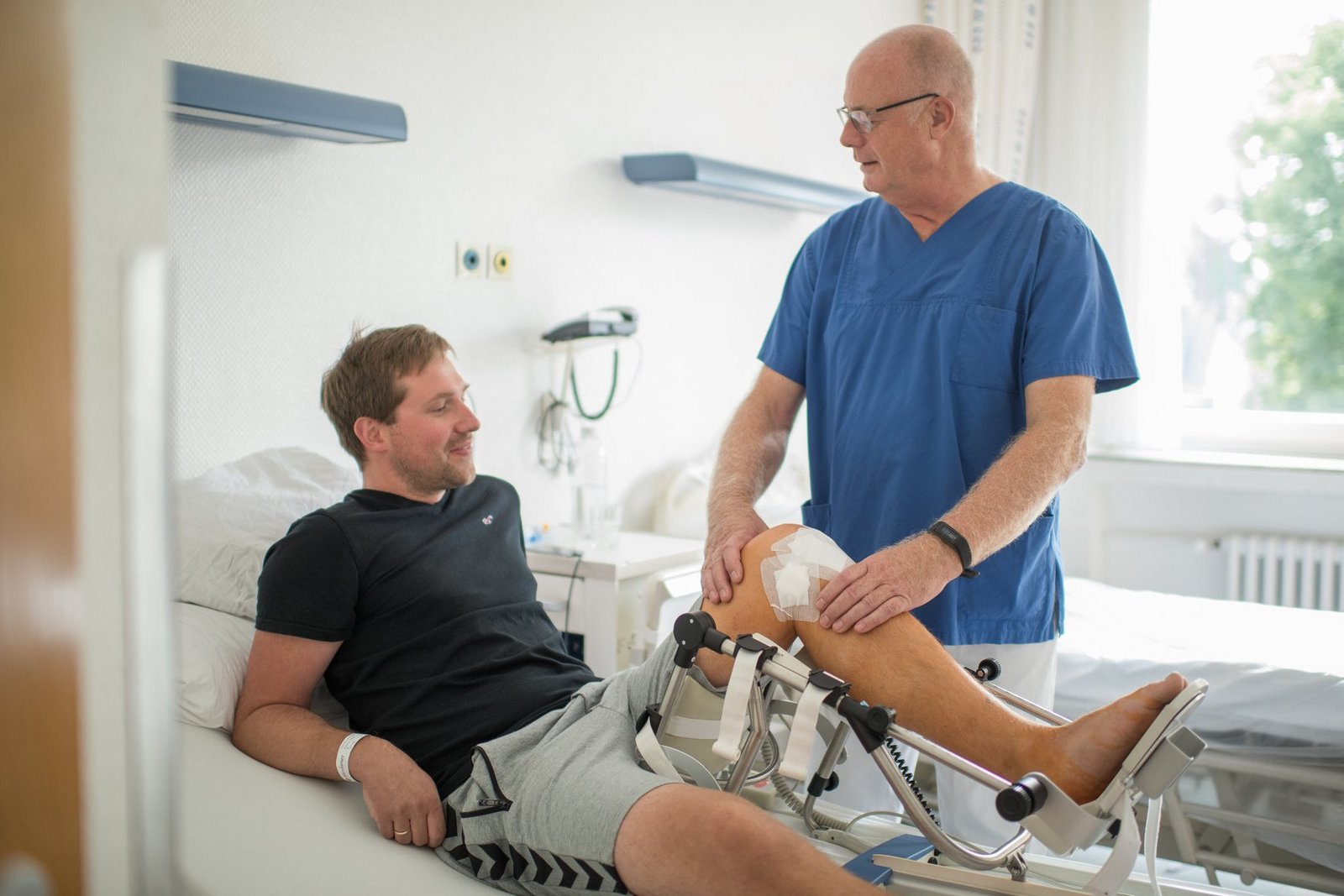 Nachuntersuchung nach Knie-OP durch Arzt am Patientenbett. Arzt tastet Knie ab.