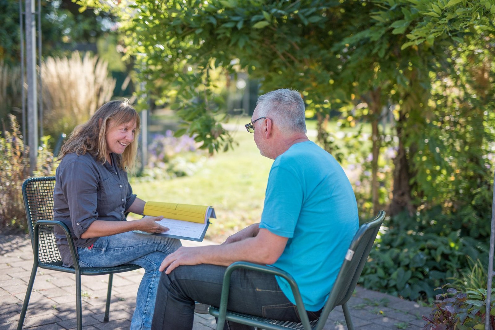 Freundlich zugewandte Krankenschwester im Pflegegespräch mit einem Patienten. Beide Personen sitzen in einer Sitzecke im Park